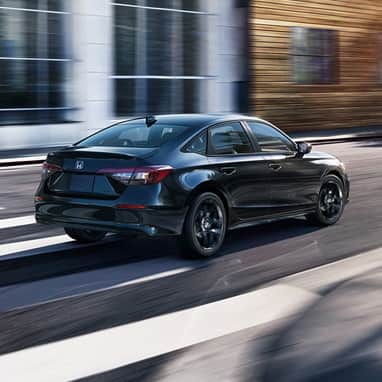2027 Honda Civic Sedan Sport Hybrid in Crystal Black Pearl rear three-quarter view driving on a city street (Compact Sedan)